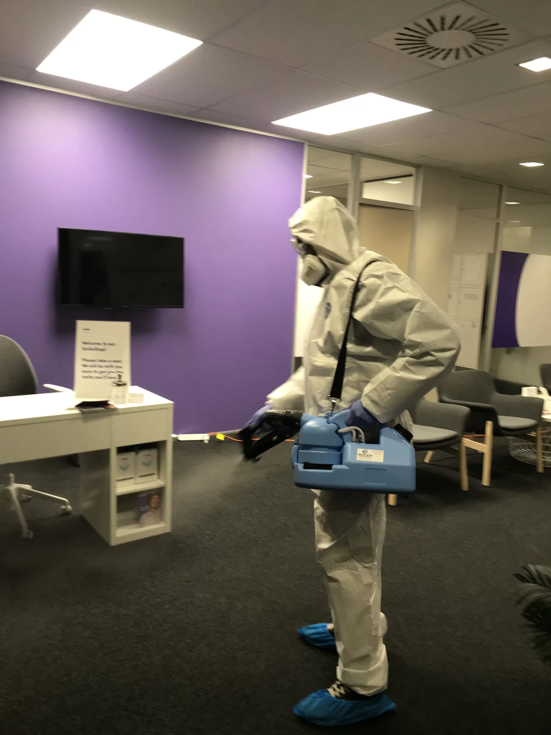 A professional cleaner in full protective gear performing fogging disinfecting services in a commercial office space.