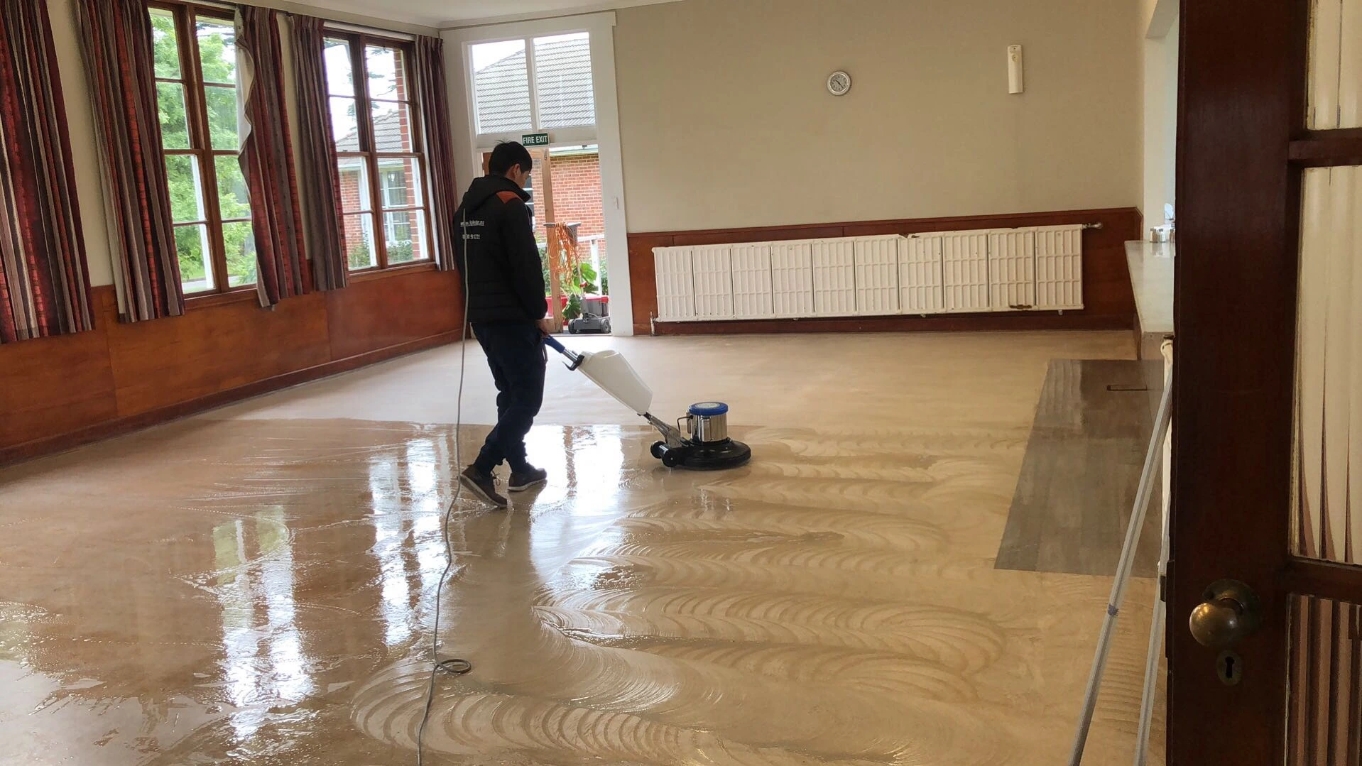 A professional cleaner using an industrial floor scrubbing machine to clean and polish a large hard floor surface in a commercial space.